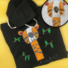Load image into Gallery viewer, black hoodie with cream drawstrings and custom embroidery of kids drawing of very happy orange tiger with closed eyes surrounded by green leaves